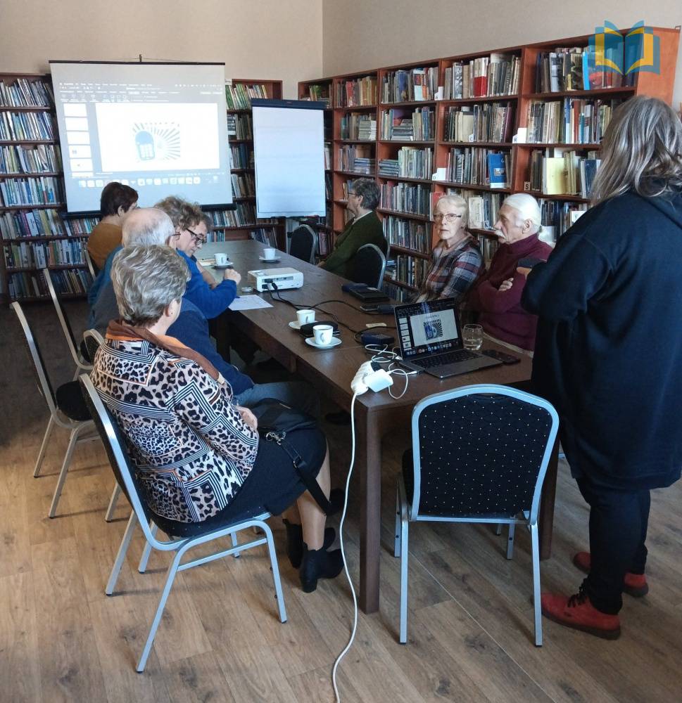 Zdjęcie: Grupa seniorów uczestnicząca w warsztatach z obsługi czytaków. W bibliotece, uczestnicy siedzą przy długim stole, patrzą na ekran, na którym jest wyświetlana prezentacja dotycząca czytaków.